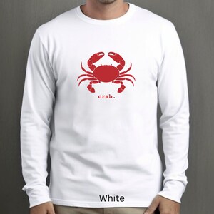 A man wearing a White unisex long sleeve t-shirt with a graphic of a red crab, above the word crab in lower case font, followed by a period.