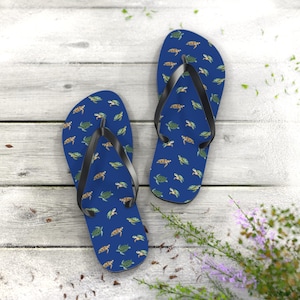 Könnte beinhalten: Zwei blaue Flip-Flops mit einem Muster aus kleinen Schildkröten in Grün und Braun. Sie haben schwarze Riemen und liegen auf einem hellen Holzhintergrund. Ein Sommeraccessoire.