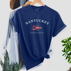 Könnte beinhalten: Ein marineblaues T-Shirt mit weißem Text, der "NANTUCKET MASSACHUSETTS" lautet, und einer Illustration eines roten Hummers.