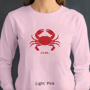 A woman wearing a Light Pink unisex long sleeve t-shirt with a graphic of a red crab, above the word crab in lower case font, followed by a period.