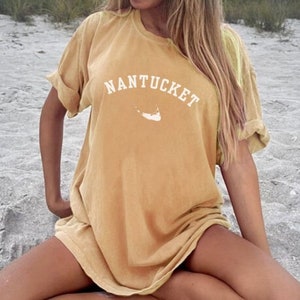 Könnte beinhalten: Eine Frau trägt ein senfgelbes T-Shirt mit dem Text "NANTUCKET" und einer weißen Umrandung der Insel Nantucket. Das Shirt hat eine lockere Passform und wirkt leicht abgenutzt.