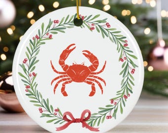 Puede incluir: Adorno navideño redondo de cerámica blanca con un diseño de cangrejo rojo. El cangrejo está rodeado por una corona verde con bayas rojas y un lazo rojo. Se adjunta una cuerda dorada. Decoración festiva navideña.