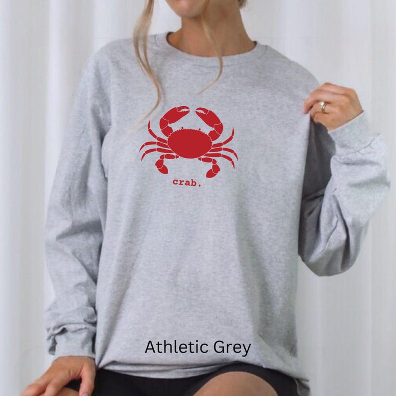 A woman wearing a Sport Grey unisex long sleeve t-shirt with a graphic of a red crab, above the word crab in lower case font, followed by a period.