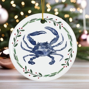 May include: A round white ceramic ornament featuring a watercolor illustration of a blue crab. The crab is surrounded by a green and red botanical wreath. A gold string is attached for hanging. The ornament is a coastal or nautical themed decoration.