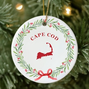May include: A white ceramic Christmas ornament with a green wreath and red berries. The ornament features a red outline of Cape Cod and the text "CAPE COD" in red lettering. A red ribbon with a bow is at the bottom of the ornament.