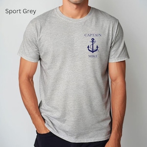 May include: Sport grey t-shirt with a crew neck. The shirt features a navy blue anchor graphic with the words "CAPTAIN" above and "MIKE" below. The shirt is made of a soft, comfortable material.
