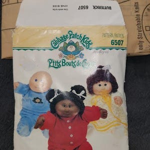 Peut inclure: Un patron de couture Butterick pour les vêtements et les habitudes des Cabbage Patch Kids. L'emballage présente des illustrations de trois poupées en tenues bleues, rouges et jaunes. Le numéro du patron est le 6507.