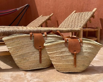 May include: Two large woven straw tote bags with brown leather handles and straps. The bags are sitting on a light brown tiled floor.