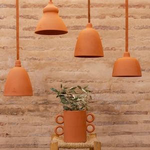 Peut inclure: Quatre lampes suspendues en terre cuite éclairent un vase en terre cuite avec des branches d'olivier. Les lampes ont un design en forme de cloche. Le vase a deux ensembles de poignées. Le fond est un mur de briques brun clair texturé.