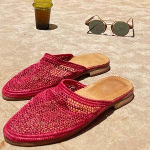 Peut inclure: Une paire de chaussons roses vifs en tissu tissé avec des semelles intérieures beiges. Des lunettes de soleil à monture dorée et verres verts sont posées à côté, ainsi qu'un petit verre de liquide. Les chaussons ont un design tissé ouvert.