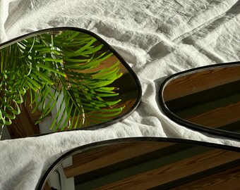 Peut inclure: Deux miroirs ovales ondulés à cadre noir reflétant une surface en bois et une plante verte.