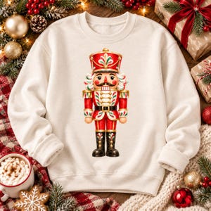 Könnte beinhalten: Ein cremefarbenes Sweatshirt mit Rundhalsausschnitt und einem zentralen Aufdruck eines Nussknacker-Soldaten. Der Nussknacker trägt eine rote Uniform mit goldenen Akzenten, einen hohen Hut und schwarze Stiefel, verziert mit grün-weißen Blattmustern.