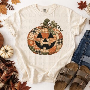 May include: A cream-colored short-sleeve t-shirt featuring a large graphic of a jack-o'-lantern pumpkin. The pumpkin design is composed of various patchwork fabric patterns in shades of orange, green, black, and white, with a stitched appearance.