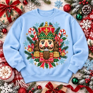 Puede incluir: Una sudadera de cuello redondo en color azul claro con un gráfico central de un soldado cascanueces rodeado de ramas de pino, bastones de caramelo, lazos rojos, campanas doradas y copos de nieve, con sombrero rojo.
