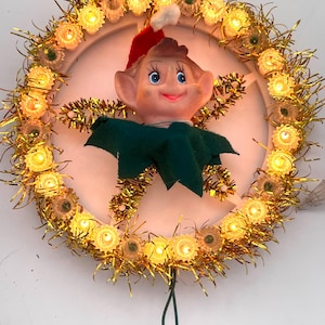 May include: A circular light-up decoration featuring a central elf figure with a red hat and green collar. The elf is surrounded by a ring of gold tinsel and small, glowing warm-white light bulbs. A green electrical cord extends from the bottom.