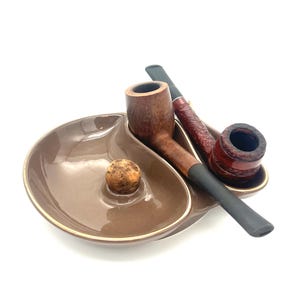 May include: A brown ceramic ashtray with a gold rim holds two tobacco pipes and a wooden ball. The pipes are made of wood and have black mouthpieces. The ashtray is a unique shape.