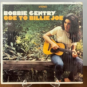 Könnte beinhalten: Albumcover für Bobbie Gentrys "Ode to Billie Joe" in einer Plastikhülle. Das Albumcover zeigt eine Frau mit langen dunklen Haaren, die eine gelbe Akustikgitarre spielt. Der Albumtitel steht in großen weißen und orangefarbenen Buchstaben.
