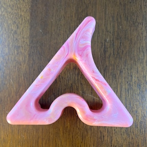 May include: A pink, marbled, triangular massage tool with a curved base. The tool has a smooth, polished finish and a hole in the center. The color palette includes shades of pink, white, and yellow. The tool is designed for body massage.