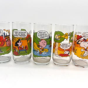 May include: A set of five clear glass tumblers featuring colorful Peanuts comic strip illustrations. Each glass displays a unique scene with characters like Charlie Brown, Lucy, and Snoopy, accompanied by text bubbles including phrases like The struggle for security is no picnic and Civilization is overrated.