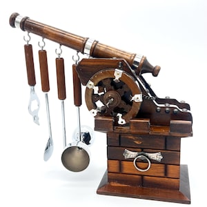 May include: A wooden bar tool set shaped like a cannon. The cannon has a dark brown finish with silver accents and includes a bottle opener, spoon, and other bar tools. The set is designed to resemble a miniature cannon.