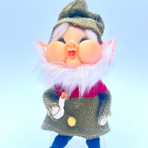 May include: A small elf figurine with a peach-colored face, pointed ears, and a white synthetic beard. The elf wears a textured olive green burlap hat and tunic, a red collar, a single yellow button, and dark blue pointed shoes, holding a small candle.