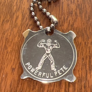 May include: A silver-colored metal keychain tag with a ball chain. The tag features an embossed image of a flexing figure and the words "POWERFUL PETE" in a circular design. The background is a wooden surface.