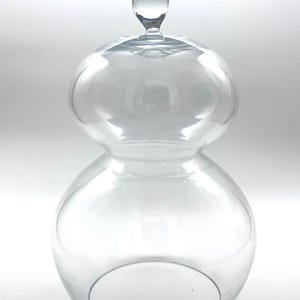 May include: A clear glass cloche with a double-bulb silhouette and a small spherical handle on top. The vessel features a wide, rounded base that tapers into a narrower middle section, topped with a smooth, rounded dome and a solid glass finial.