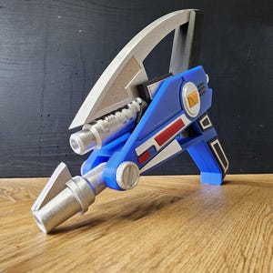 May include: A blue and silver prop weapon with a futuristic design. The prop features a silver blade, a blue body with red and white accents, and a silver barrel. The weapon is likely a cosplay or display item.