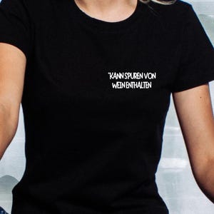 Könnte beinhalten: Schwarzes T-Shirt mit dem weißen Text "KANN SPUREN VON WEIN ENTHALTEN" auf der oberen linken Brust. Das Shirt ist kurzärmlig und aus weichem Material. Der Hintergrund ist hell und verschwommen.