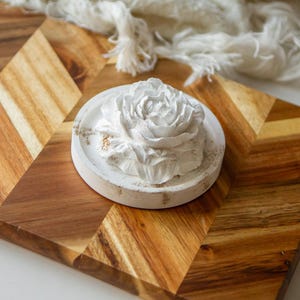 May include: A white, sculpted flower-shaped object sits atop a circular, off-white concrete tray. The tray features subtle, light brown textured markings along its edge, providing a contrast to the smooth, layered petals of the floral sculpture.