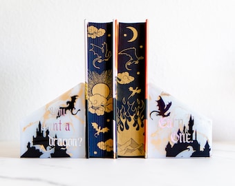 May include: Decorative bookends with a fantasy theme. The bookends are white with gold marbling and feature a black silhouette of a castle and dragon. The books have a navy blue and gold design with dragons, clouds, and the sun. The text on the bookends reads "Want a dragon?" and "Earn One!"