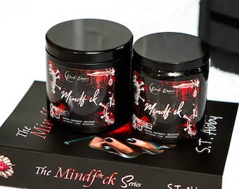 May include: Two black candle jars with red and white labels. The labels feature the text "Mindf*ck" and "Dark Desire Candles". The candles are sitting on a black book with the text "The Mindf*ck Series" printed on the cover.