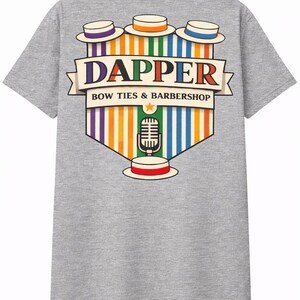 May include: Gray t-shirt with a colorful graphic featuring the word "DAPPER" above "BOW TIES & BARBERSHOP." The design includes bowler hats, a microphone, and a star, with vertical stripes in various colors.