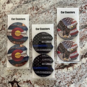 May include: Set of six car coasters featuring different designs. Two coasters feature a wooden background with the Colorado state flag, two coasters feature a black and white American flag with a blue stripe, and two coasters feature an American flag with a bald eagle.