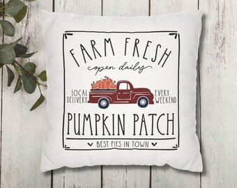 May include: White square pillow with a black framed design. The design includes the text "FARM FRESH open daily" above a maroon pickup truck filled with pumpkins. Below the truck is the text "PUMPKIN PATCH best pies in town".