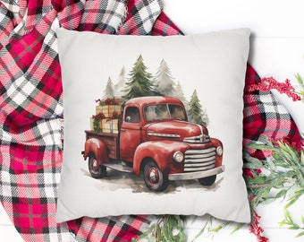 May include: A decorative square pillow featuring a watercolor painting of a vintage red pickup truck carrying wrapped gifts. The truck is set against a backdrop of evergreen trees. The pillow has a white background and is surrounded by red and black plaid fabric.
