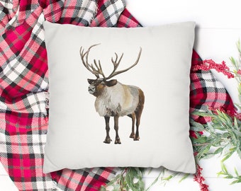 May include: A white throw pillow with a watercolor illustration of a reindeer with large antlers. The pillow is on a white surface with a red and white plaid blanket and green foliage.