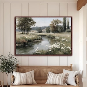 May include: A rectangular landscape painting in a dark brown frame depicting a calm river flowing through a grassy meadow with white wildflowers, scattered trees, and a soft, muted sky in shades of green, brown, and cream.