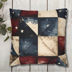 May include: Square throw pillow with a patchwork design in patriotic colors. The pillow features a pattern of red, white, and blue squares with white stars, evoking a vintage American flag aesthetic. The pillow is placed on a white wooden surface.