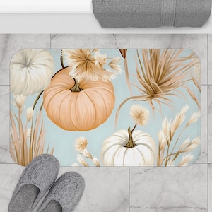 May include: A light blue bath mat with a fall theme featuring white and orange pumpkins, dried grasses, and fluffy white flowers.