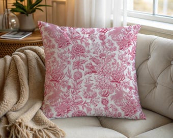 May include: A square decorative pillow with a white background and a pink floral toile pattern. The pillow is on a beige sofa, with a tan throw blanket visible. The pillow's design features flowers and foliage.