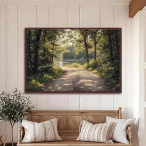 May include: A framed landscape painting depicts a sunlit dirt road winding through a forest towards a body of water. The artwork features lush green trees, dappled sunlight, and a warm, inviting color palette. The frame is a dark brown color.