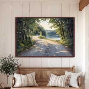 May include: A framed landscape painting depicts a winding dirt road through a forest, leading to a body of water. The artwork features a palette of greens, browns, and blues, with sunlight filtering through the trees. The painting is displayed above a wooden bench with striped pillows.