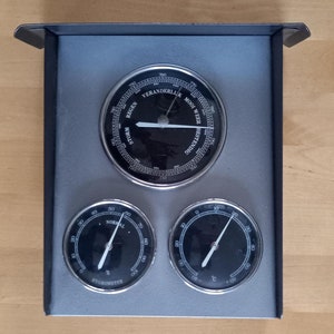 May include: A silver-toned weather station with three round gauges. The top gauge is a barometer with the text "VERANDERLUK MOOI WEER BESTENDIG" and markings for "STORM", "REGEN", and "7150". The left gauge is a hygrometer with the text "NORMAL" and markings for "%" and "HYGROMETER". The right gauge is a thermometer with markings for "C" and "00".