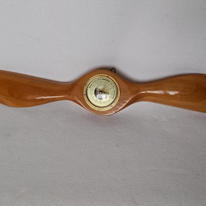 May include: A wooden aeroplane propeller with a round barometer in the centre. The propeller is light brown and the barometer is brass with a white face.