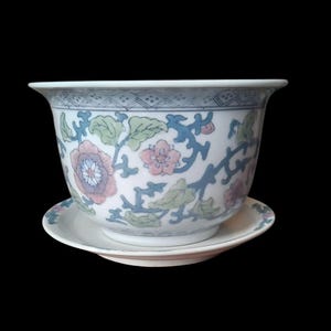 Pode incluir: Uma taça de cerâmica branca com pires a condizer, apresentando um padrão floral delicado com flores rosa suave, folhas verde-claras e videiras azuis. O rebordo da taça é decorado com um padrão geométrico subtil de losangos em azul.
