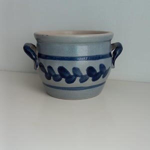 Op de afbeelding: Een grijze keramische pot met blauwe beschilderde ontwerpen en twee handvatten. De pot is rond en heeft een brede opening.