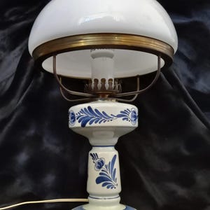 Może przedstawiać: Biała ceramiczna lampa stołowa z niebiesko-białym wzorem kwiatowym. Lampa ma biały szklany klosz z falistym brzegiem i mosiężną ozdobą.