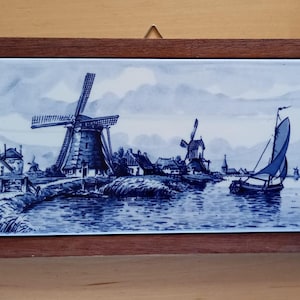 Può includere: Una piastrella di Delft blu e bianca incorniciata che rappresenta una scena di mulino a vento olandese con due mulini a vento e una barca a vela su un canale. La piastrella è incorniciata in una cornice di legno scuro.