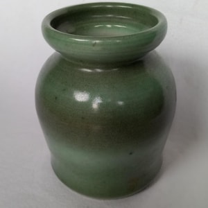 Può includere: Un piccolo vaso in ceramica verde con corpo arrotondato e un labbro leggermente svasato. Il vaso presenta un effetto sfumato sottile, passando da un verde più scuro nella parte inferiore a un verde più chiaro nella parte superiore.
